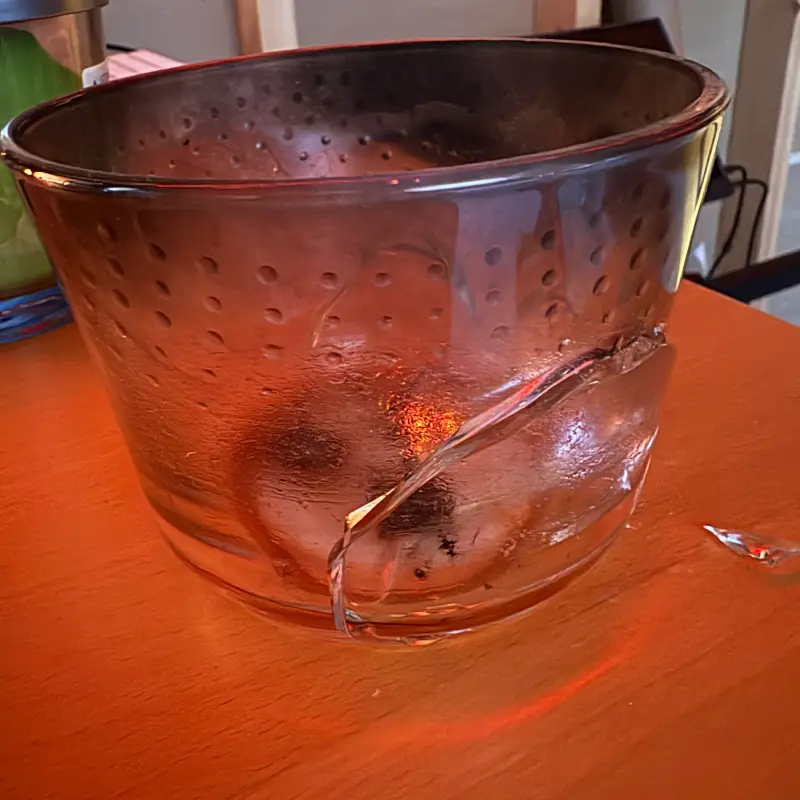 A dimpled clear glass bowl on an orange wooden table, cracked from rim to base with a shard broken away beside it
