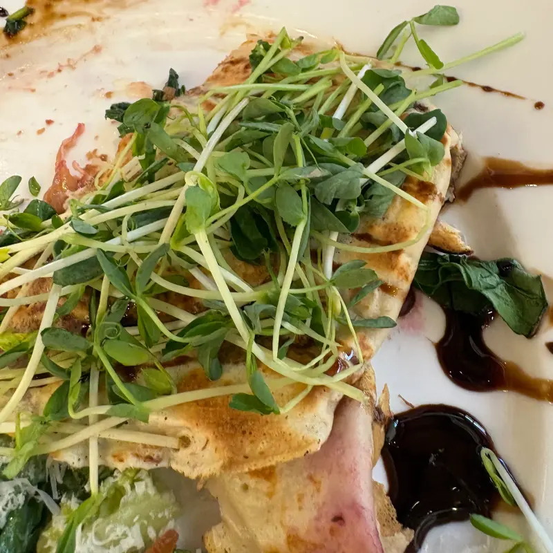 A part-eaten flatbread on a white plate under a heap of pea shoots, with prosciutto at its edges and dark balsamic pooled alongside