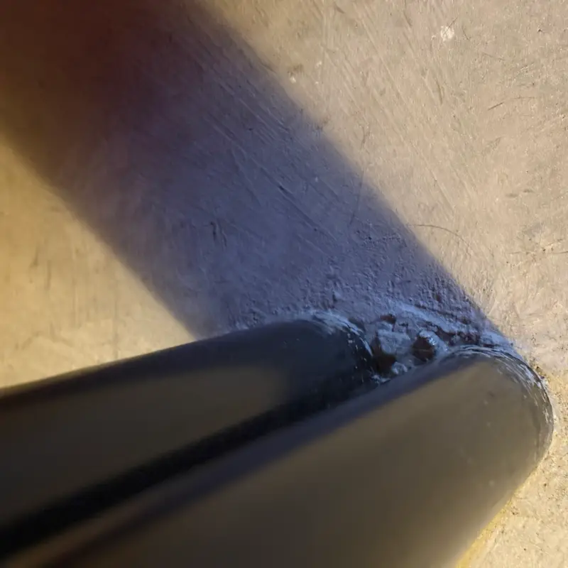 The cut end of a dark metal tube against a pale scratched plaster surface, crusted with pale debris where they meet, its shadow thrown hard across the wall by low sunlight