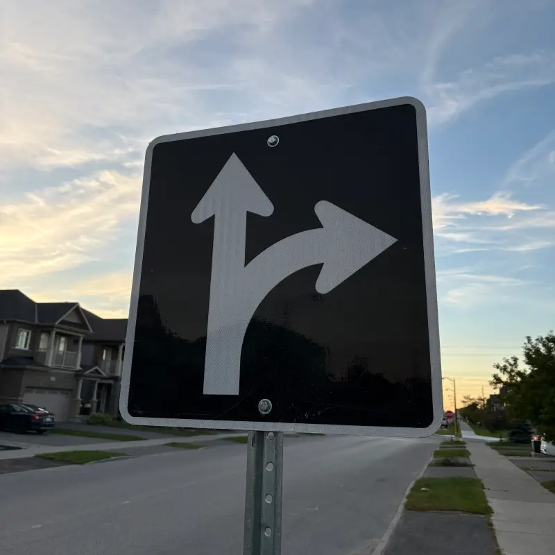 A square black road sign with white straight-ahead and right-turn arrows, bolted to a galvanized post and shot from close and below so it leans back against a pale evening sky, with suburban brick houses and a parked car at the left and empty asphalt and sidewalk running away to the right