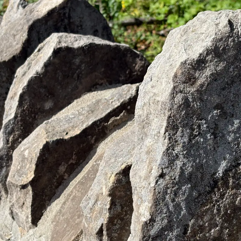 Broken grey limestone blocks stacked against one another in hard overhead sun, the fractured faces bright and the wedge-shaped gaps between them dropping to black, with a band of green foliage along the top edge